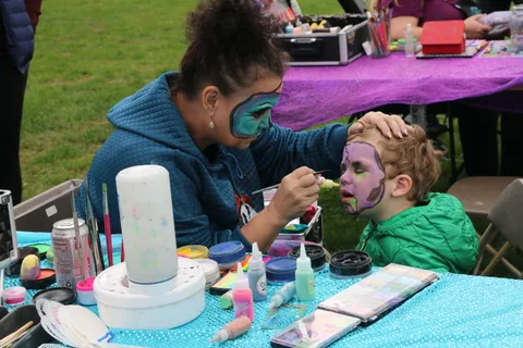 A face painter carefully paints a young boy's face with blue and black designs.