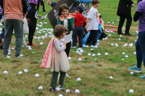 A child in costume stands amid other kids and scattered balls on a grassy field.