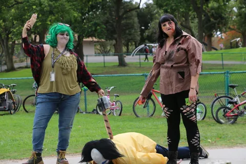 Performers pose beside a fallen costumed figure on the grass.