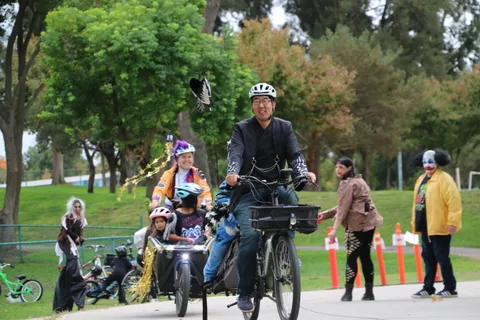 A group of adults and children ride bikes along a park path, one towing a straw-filled cart.