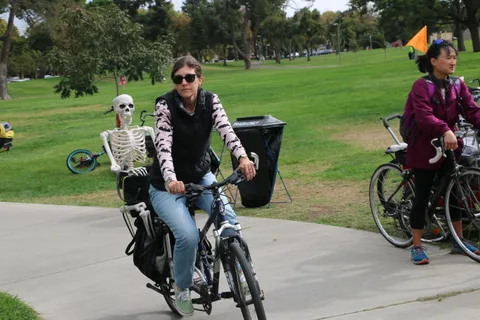 A cyclist in a skeleton-print jacket pedals past another rider in the park.