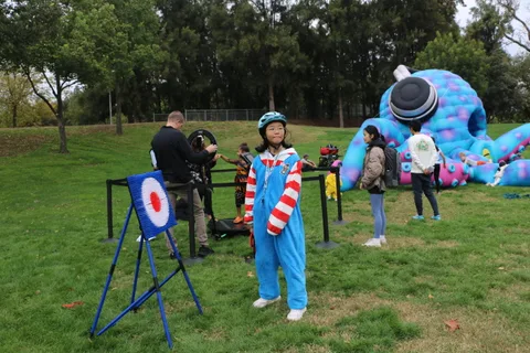 A child in a cat costume stands at an archery range beside a giant inflatable creature.