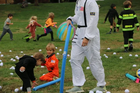 An adult in a white astronaut costume stands in a field as children in orange jumpsuits play nearby.