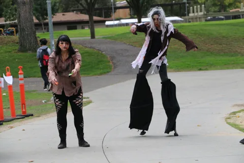 A performer in tattered zombie costume walks along the path as a stilt walker follows.