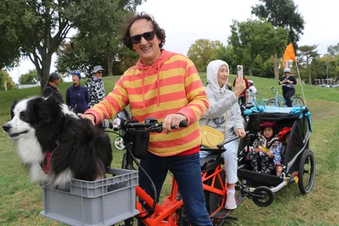 A man in a striped shirt with a dog in a bike crate rides alongside a costumed companion.