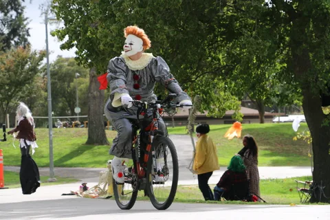A man in white clown face paint and an orange wig rides a decorated bicycle through the park.