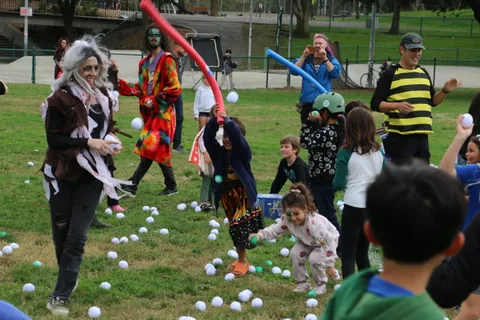 Costumed adults play with kids using a red foam sword amid scattered balls.