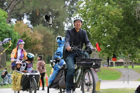 A man pedals a cargo bike with children aboard as a girl walks alongside on the path.