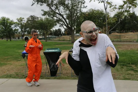 Two people in bald-cap zombie costumes lunge toward the camera with dramatic screaming expressions.