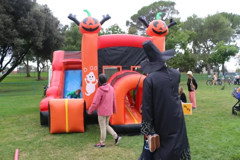 A child plays on an orange pumpkin-themed inflatable slide as a hooded figure walks by.