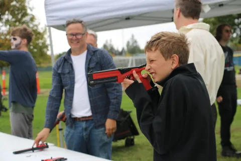 A man and a boy stand together under a tent, one holding a red toy blaster.