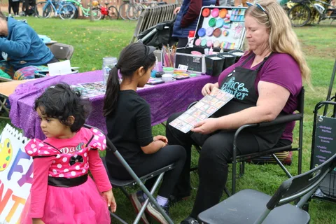 A face painter reviews her supplies while a toddler looks on nearby.