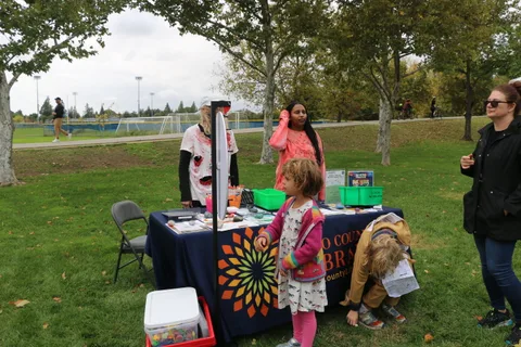 Children and staff gather around booths in a park, with a colorful prize wheel in the distance.