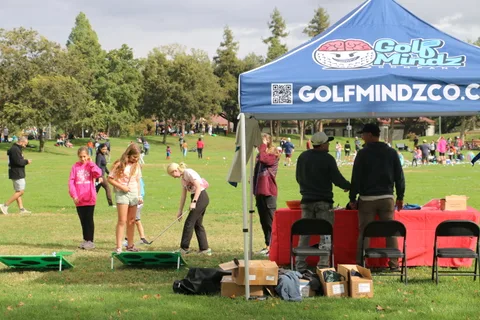 Children play cornhole games at a GolfMindz tent booth in the park.