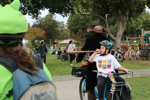 A costumed rider pulls a bike carrying a child blowing a horn through the park.