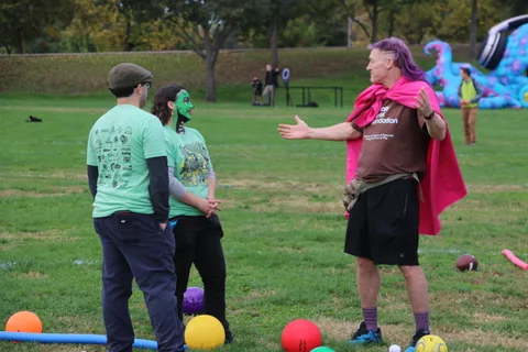 Two men in green shirts stand talking near colorful balls scattered on the grass.