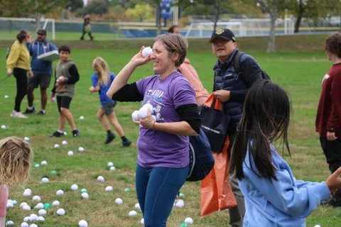 A woman in a purple shirt tosses a white ball while children play among scattered balls on the grass.