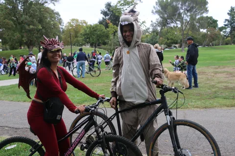 A woman in a red costume and a man in a hooded costume pose with their bikes and a dog.