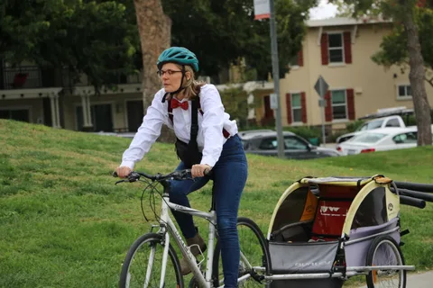 A woman with glasses and a bow tie rides a bike pulling a cart trailer.