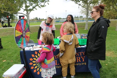 Children gather at a colorful prize wheel booth in the park.