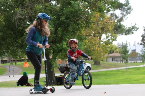 A mother and son wearing helmets ride a scooter and bike together in the park.