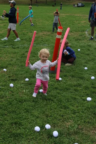 A toddler strides across the grass holding a striped foam pole in each hand.