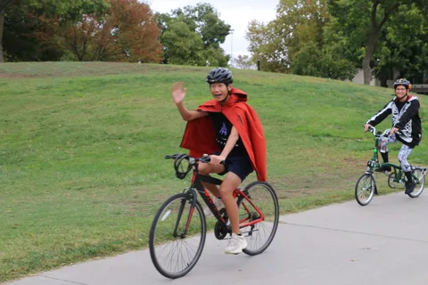 A child in a red cape waves while riding a bike.