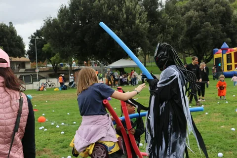 A girl rides a pool noodle like a broomstick beside a hooded, robe-clad figure.