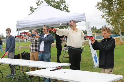 A group of men aim red toy blasters at each other across a table under a tent.