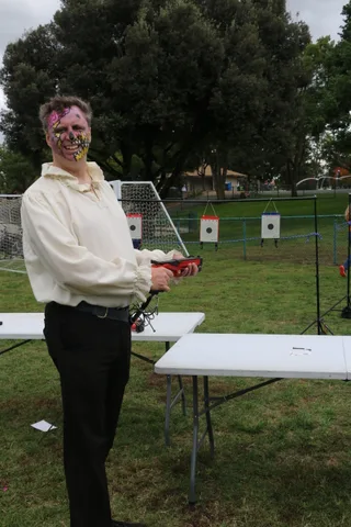 A person in a cream blouse aims a red toy blaster down an outdoor range.