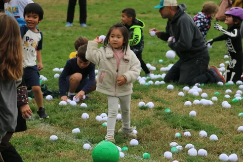 A girl in a cream jacket crouches to pick up a ball, focused and smiling.