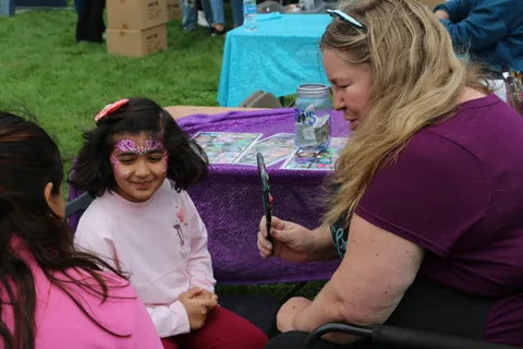 A child with pink face paint sits at a face painting booth in the park.