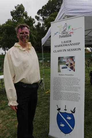 A man in detailed zombie face paint poses beside a Davis Pentathlon sign under a tent.