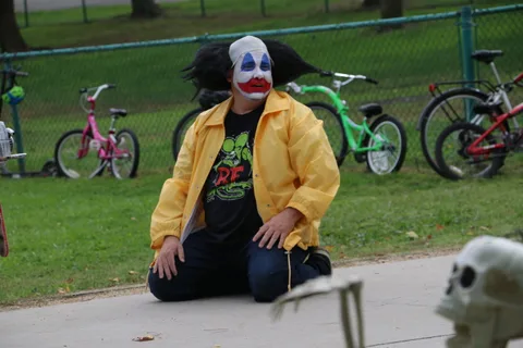 A performer in green wig and yellow jacket clown makeup kneels for the camera.