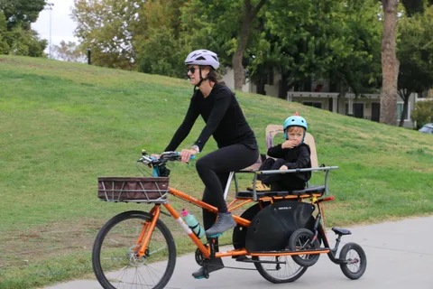 A woman rides an orange cargo bike with a child seated behind her.
