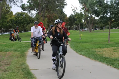 Two riders in plaid pants pedal toward the camera on a tree-lined path.