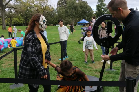 A woman in skull face paint stands with a girl in a butterfly costume as a man adjusts a camera rig.