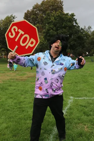 A man in a patterned costume gestures with arms wide in the grass.