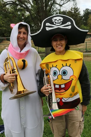 Two women in cartoon sponge costumes laugh together with their trumpets.