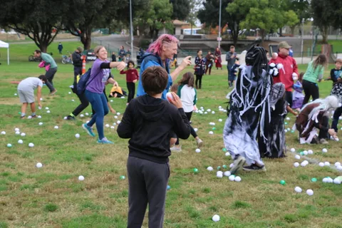 A ghost-costumed figure stands among a scattered crowd on the grass.