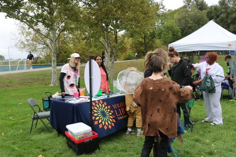 A staff member in skull face paint talks with children at a library booth.