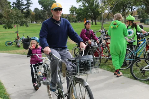 A man rides his bike with a child in a helmet in the cargo basket as costumed riders pass.