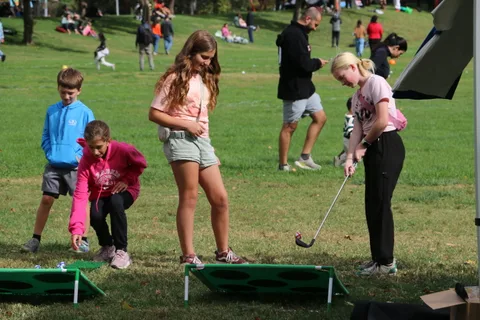 A girl swings a golf club during a cornhole game as an instructor and friend look on.
