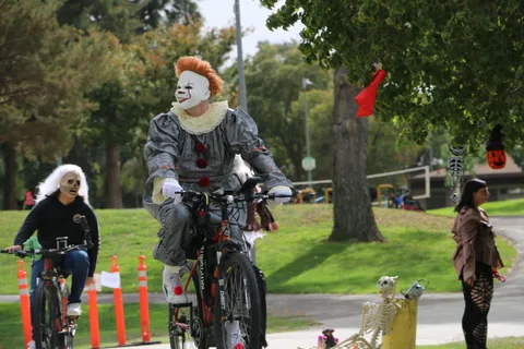 A man in a spooky clown costume rides a decorated bicycle down a park path.