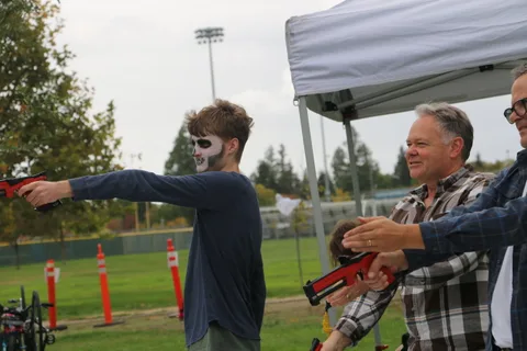 A teenager in skull face paint aims a toy blaster at a man reaching toward him.