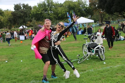 A man in a pink cape and a woman in a skeleton costume interact near parked bikes.