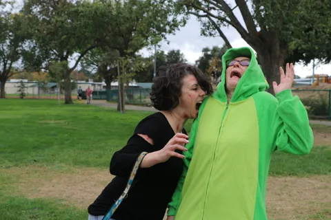 Two women, one in a green costume, embrace and smile in the park.