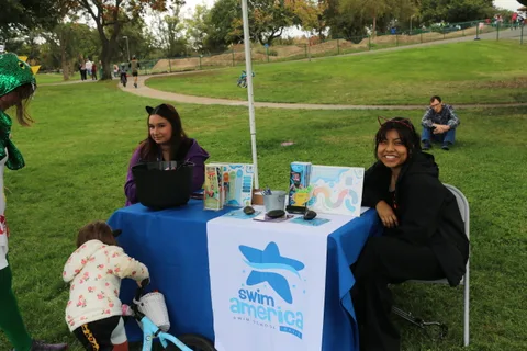 Two people sit behind a Swim America table with a child on a bike nearby.