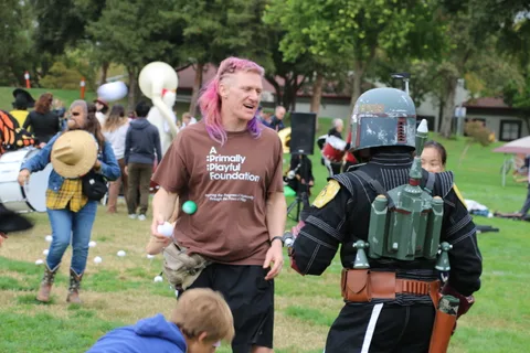 Two men in costume, one with pink hair and one wearing a full helmet, stand together at the festival.