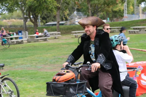 A man in a pirate hat rides a cargo bike carrying a child through the park.